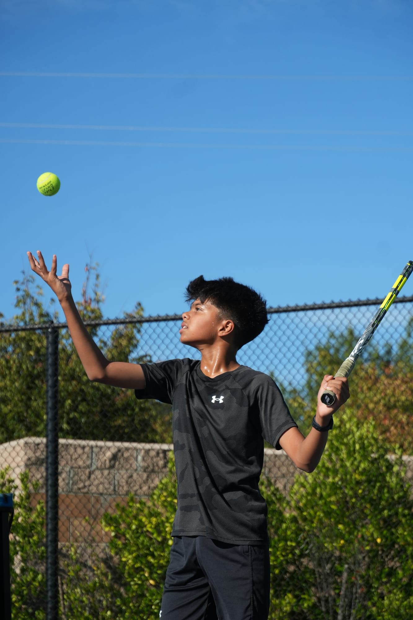 A competitive junior player tossing the ball to serve under a clear blue sky