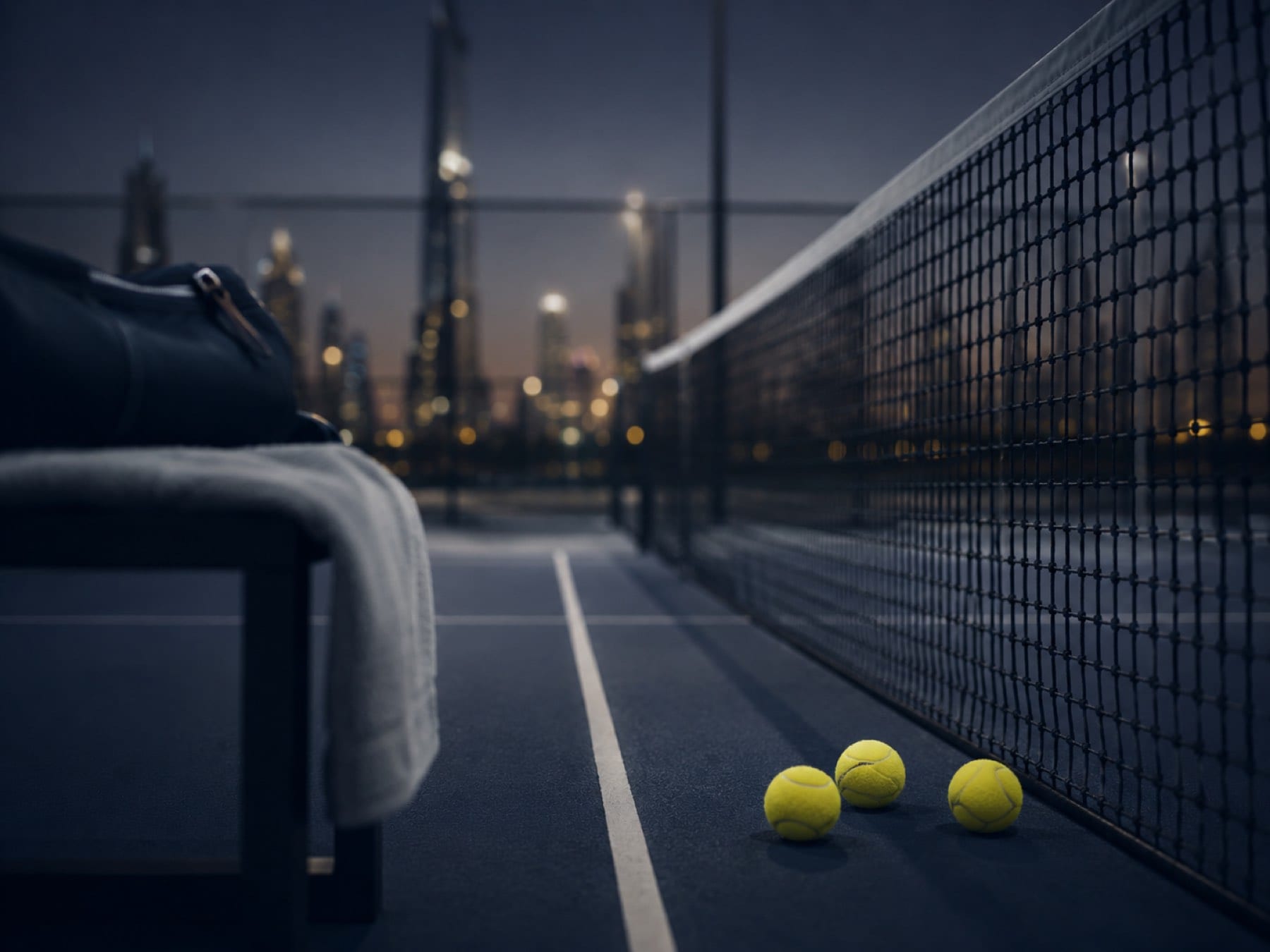 A courtside bench with a bag and tennis balls by the net, the Dubai skyline beyond